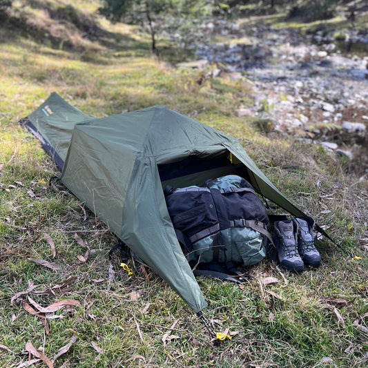 Green tent set up outdoors with camping gear inside, near a stream and trees.