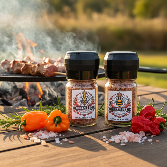 Two bottles ofQueen Bee Habanero & Reaper Salt with peppers and salt on a wooden surface with a grill in the background.